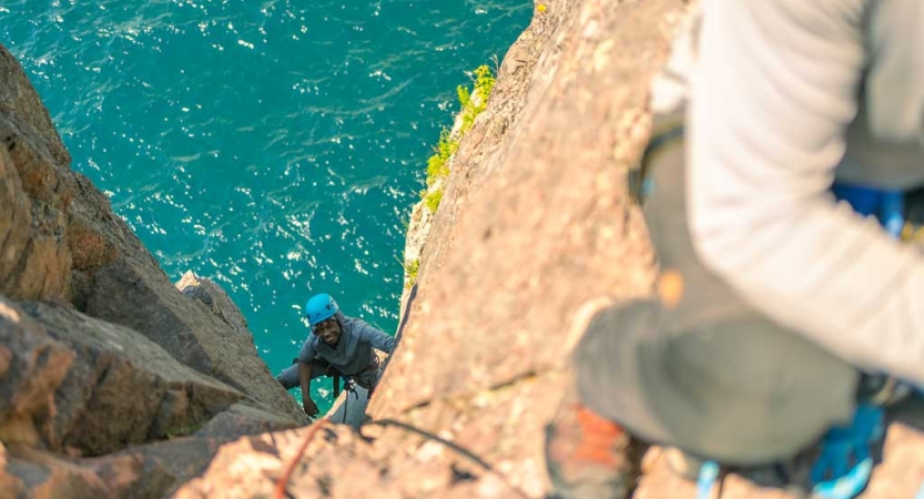 A person wearing safety gear is secured by ropes as they look up at the camera and smile while rock climbing. They are high above a blue body of water. 